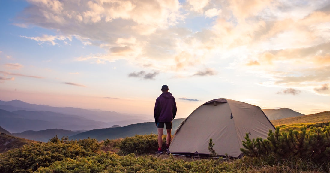 A man wearing shorts, a jacket, and a hat, standing outside of his tent. He is looking out at a mountain range.