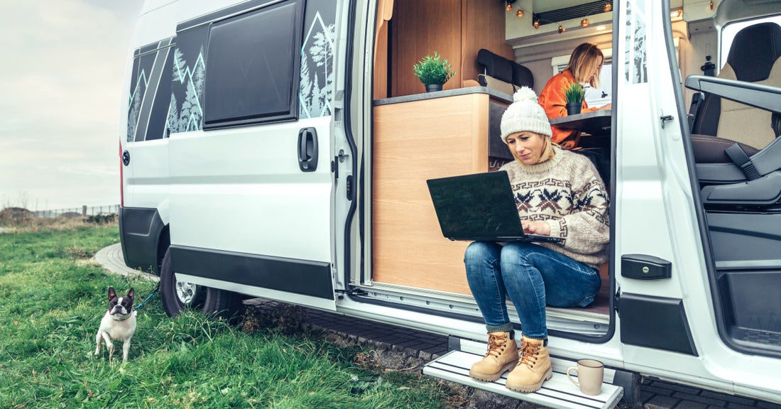 A woman sitting at the open door of a camper van with a laptop on her lap. Another woman is sitting at a table inside.