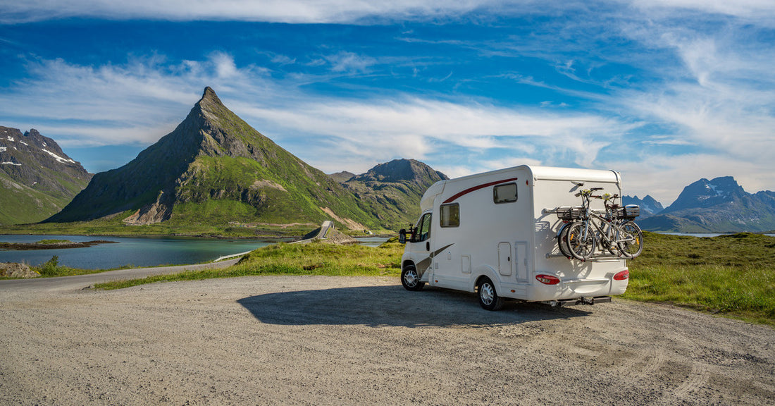 A white RV with bikes attached to the back is parked in front of a body of water and several large mountains on a sunny day.