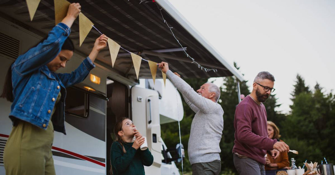 A multigenerational family prepares a cookout outside their camper. They set up their sun visor and seem to be in a wooded area.