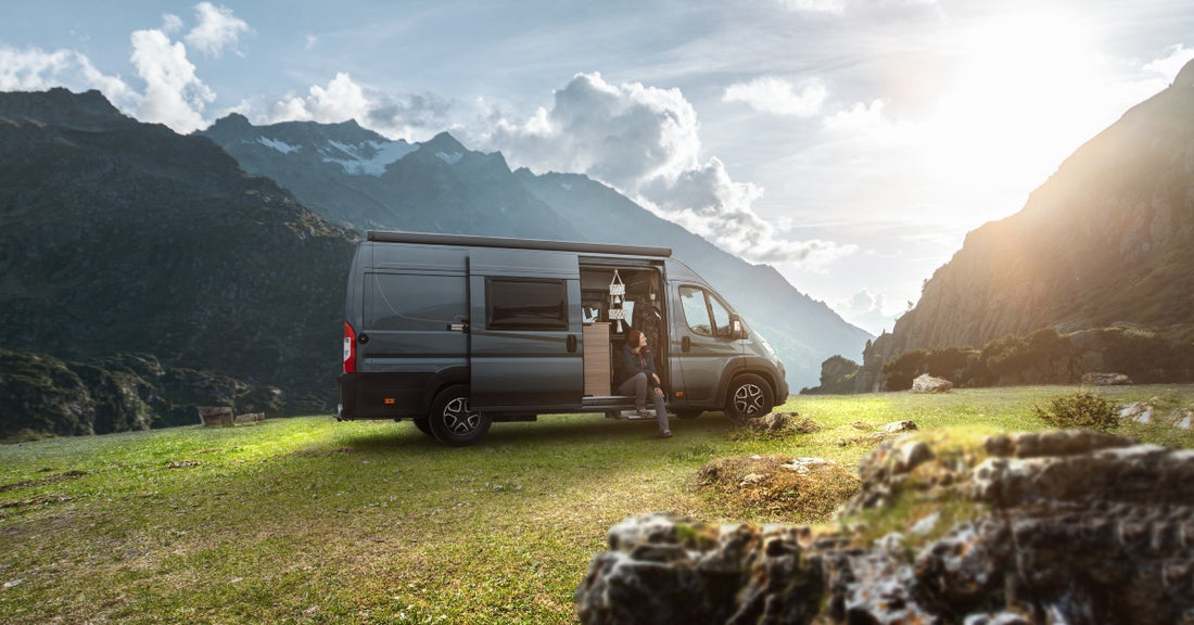 A parked camper van in a field with mountainous terrain has the sliding door open. A person sits on the ledge.