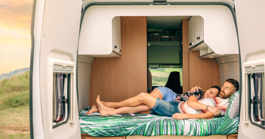 A young couple sleeping together on a bed inside a camper. The camper doors are open, and it's parked on an open field.