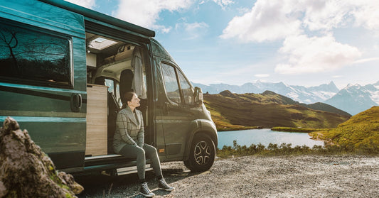 A woman sitting in the front doorway of her RV camper van. She stares at the mountainous landscape around her.