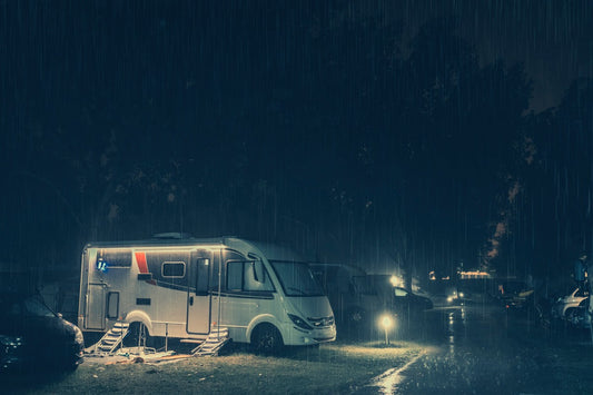 A parked RV with lights illuminates the side doors in a campsite on a rainy night. A small light shines on the road.